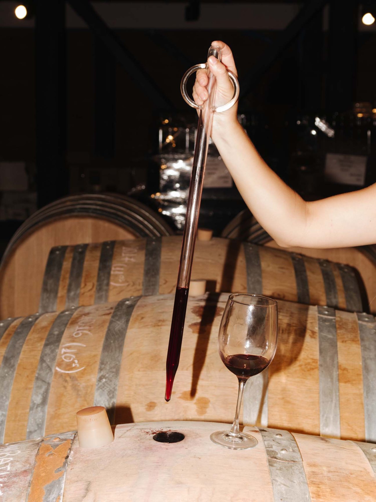 Hand holding a glass wine thief above a glass of mavrodaphne wine beside wooden wine barrels at Sclavos winery.