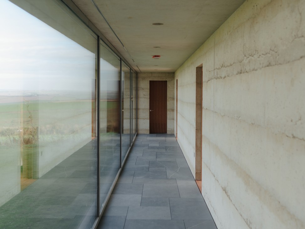 Long concrete corridor with floor-to-ceiling plate-glass windows framing Devon countryside views and brown wooden doors along the interior wall.