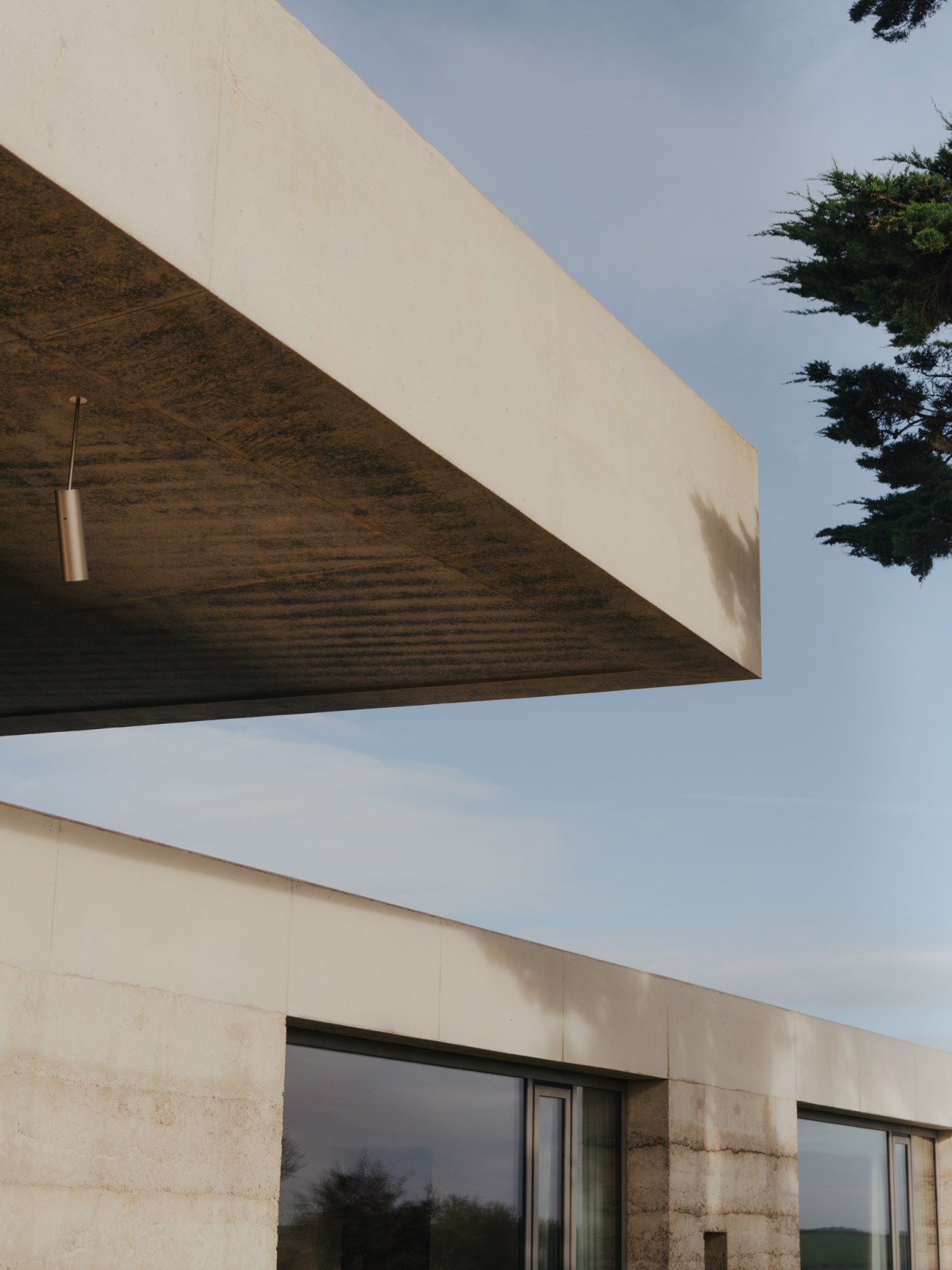 Cantilevered concrete roof overhang with wooden soffit, rammed earth walls and large windows against blue sky.