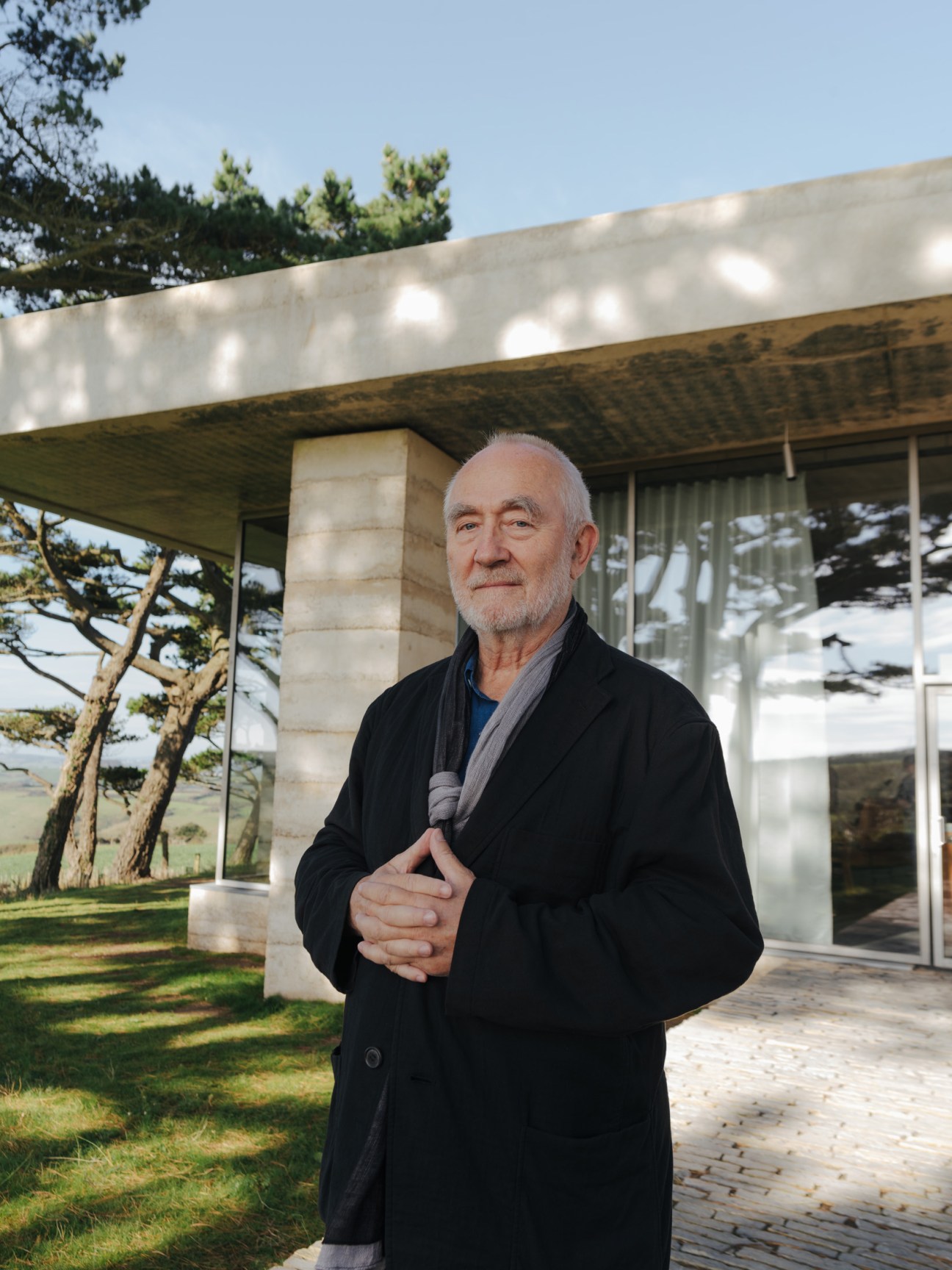Architect Peter Zumthor stands outside the Secular Retreat house in Devon, with concrete walls and glass visible behind him.