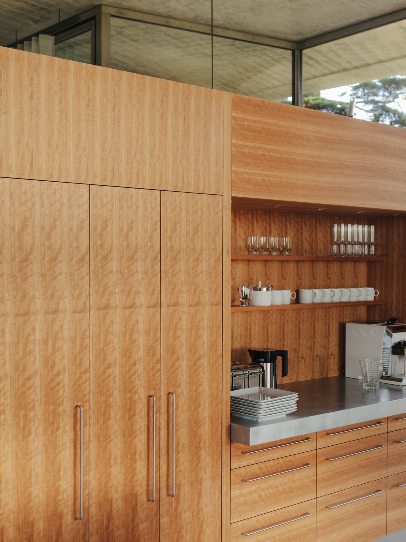 Minimalist kitchen with light wood cabinetry, open shelving displaying white dishes, and concrete ceiling in Peter Zumthor's Secular Retreat house.