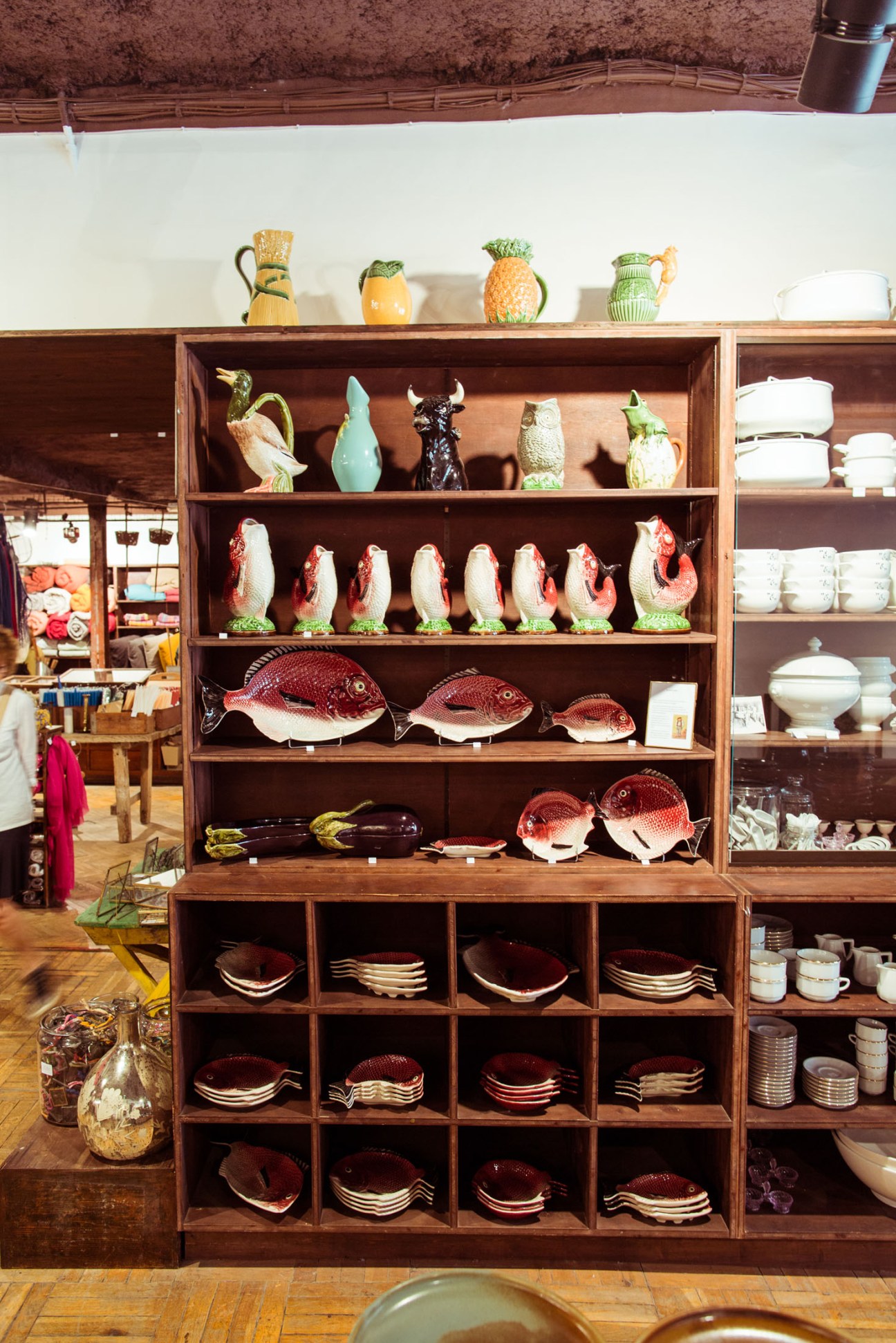 Wooden shelving unit displaying Bordallo Pinheiro ceramic pieces including fish-shaped tureens, decorative birds, and red plates.
