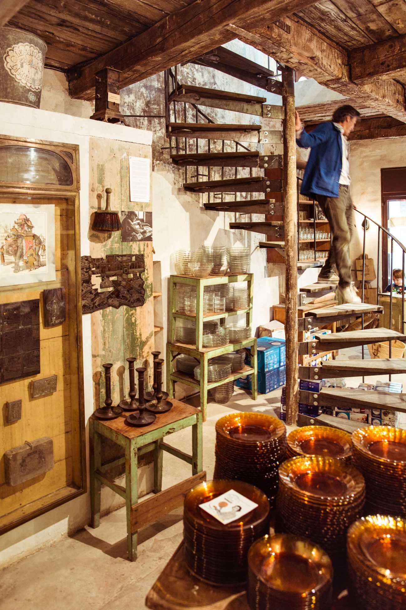 Interior of Maison Empereur showing glass tableware displays, wooden shelving, and a person climbing stairs.