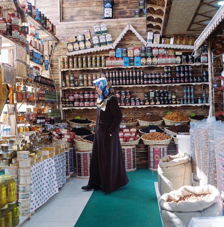 Woman in blue headscarf and black coat stands in a shop stocked with jarred goods and packaged foods on wooden shelves.