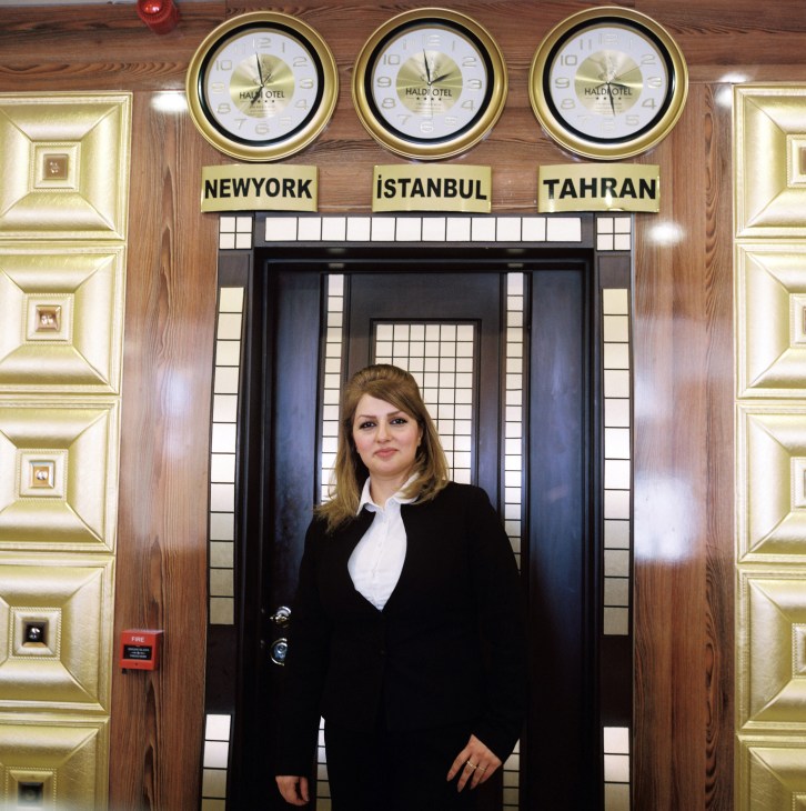 Sanaz Mahmoudi, receptionist at Haldi Hotel, stands in the marble lobby beneath clocks showing New York, Istanbul and Tehran time zones.