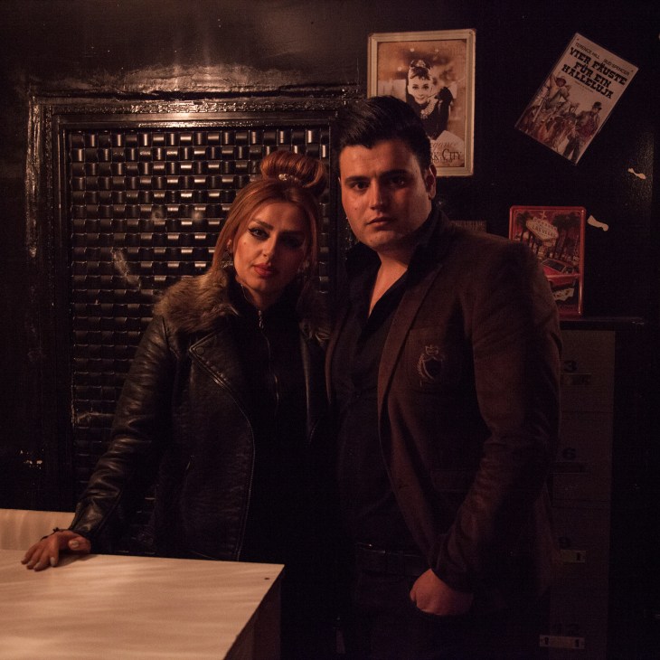 A woman and man stand in a dimly lit bar decorated with vintage movie posters and a brick fireplace.