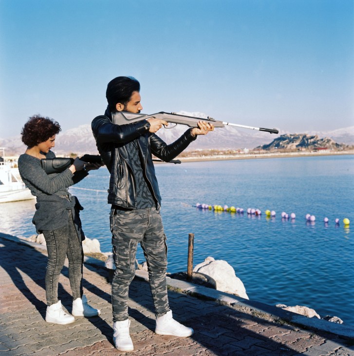 Two Iranian friends aim an air rifle at colorful balloons floating on Lake Van with snow-capped mountains in the background.