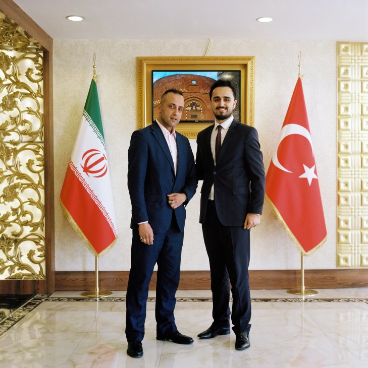 Two men in business suits shaking hands in front of Iranian and Turkish flags in an ornate lobby.