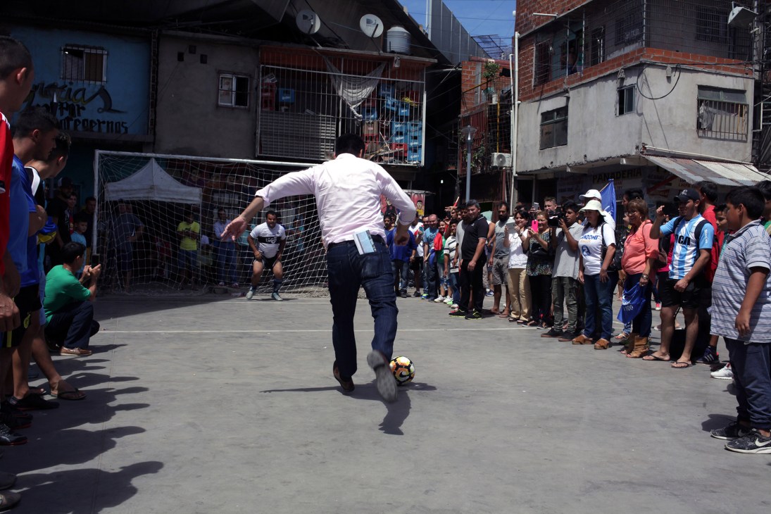 Man in white shirt kicks soccer ball in street surrounded by spectators in urban neighborhood.