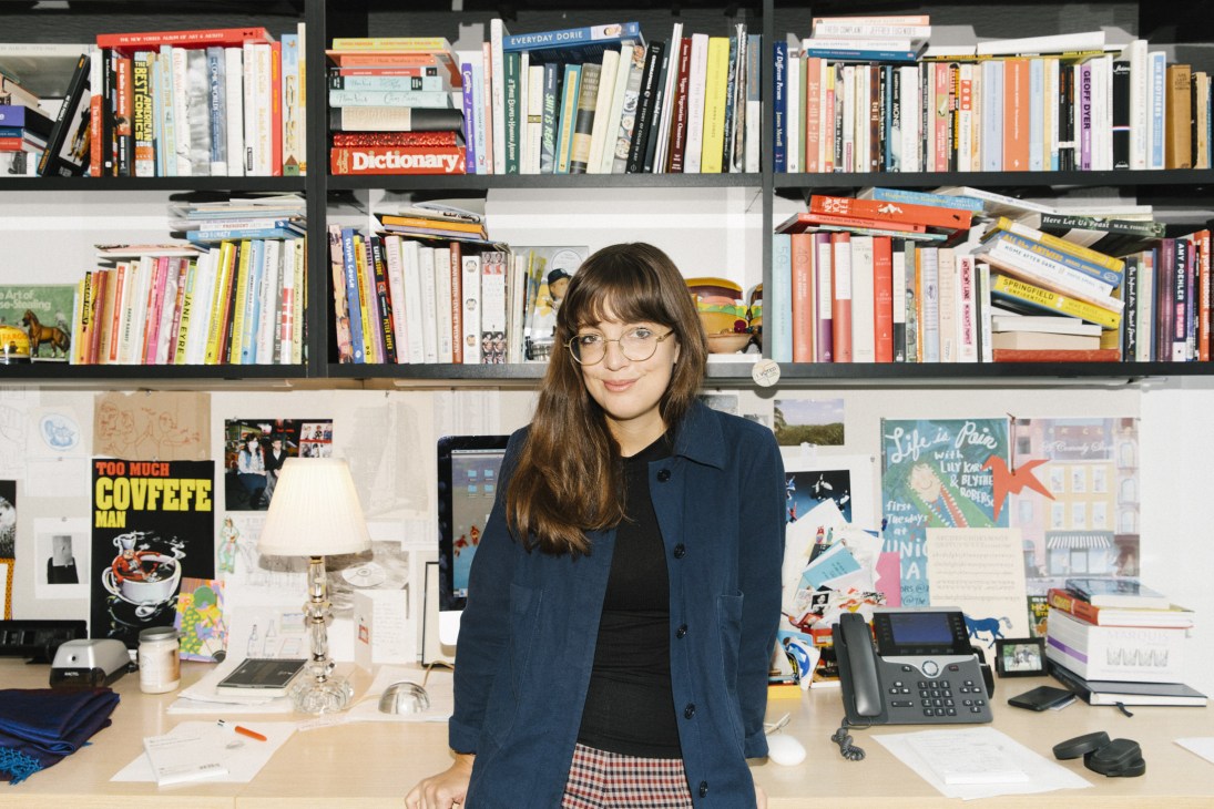 Emma Allen seated at a desk in The New Yorker offices with bookshelves and cartoon artwork behind her