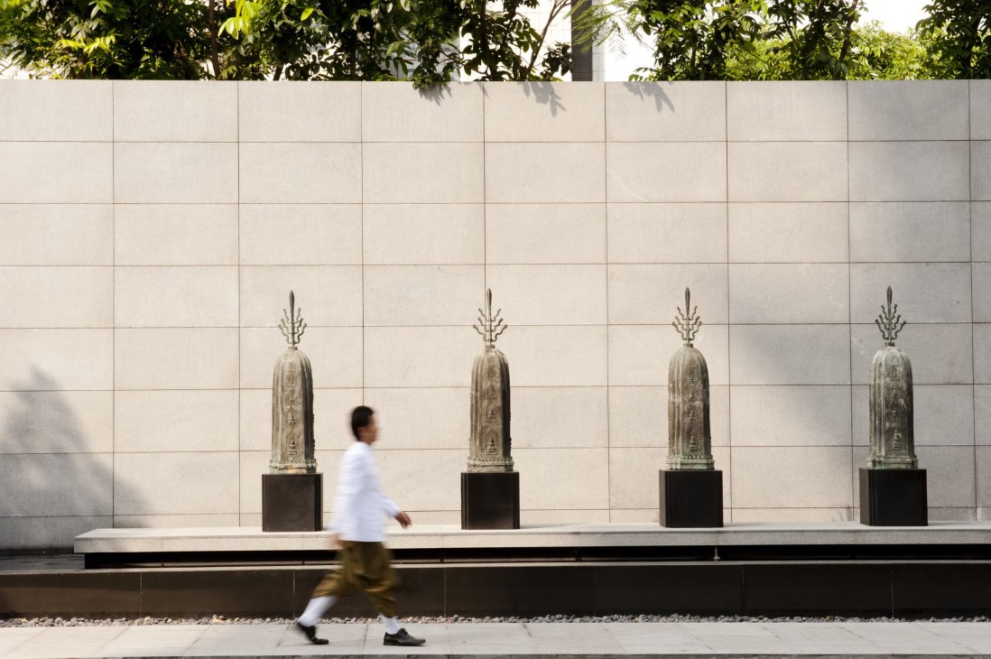 A person in white shirt jogging past the entrance of The Sukhothai hotel, with four decorative stone stupas against a minimalist tiled wall.