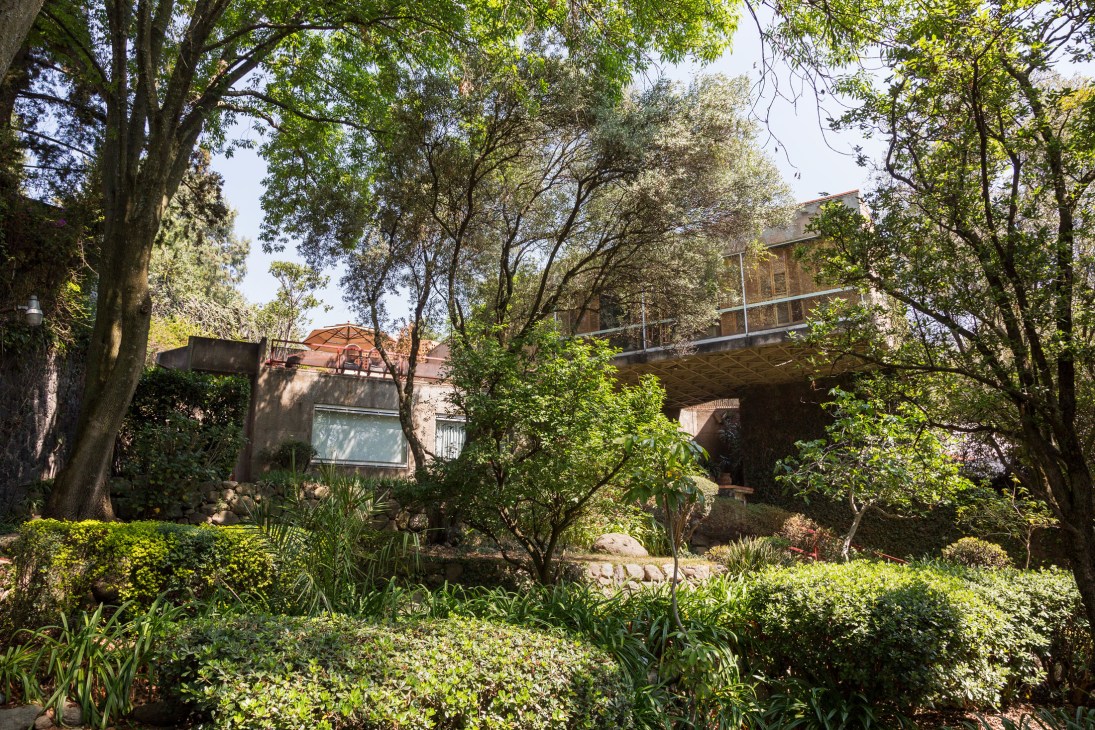 Lush garden surrounding a modernist concrete house with trees and vegetation in the foreground.