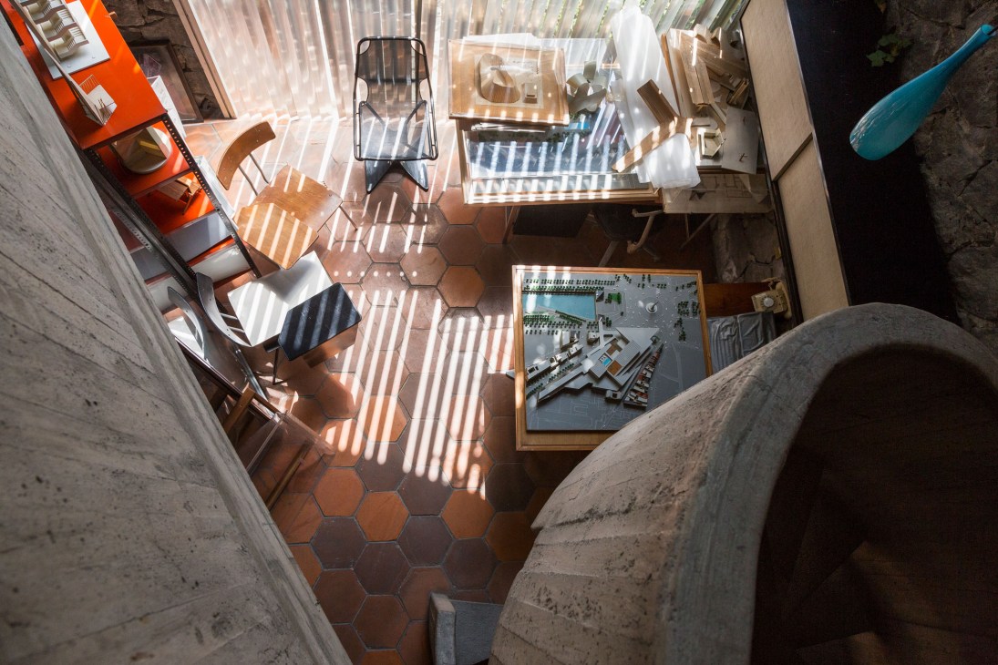 Overhead view of a workshop space with architectural models, wooden furniture, and design sketches scattered on hexagonal tile flooring.