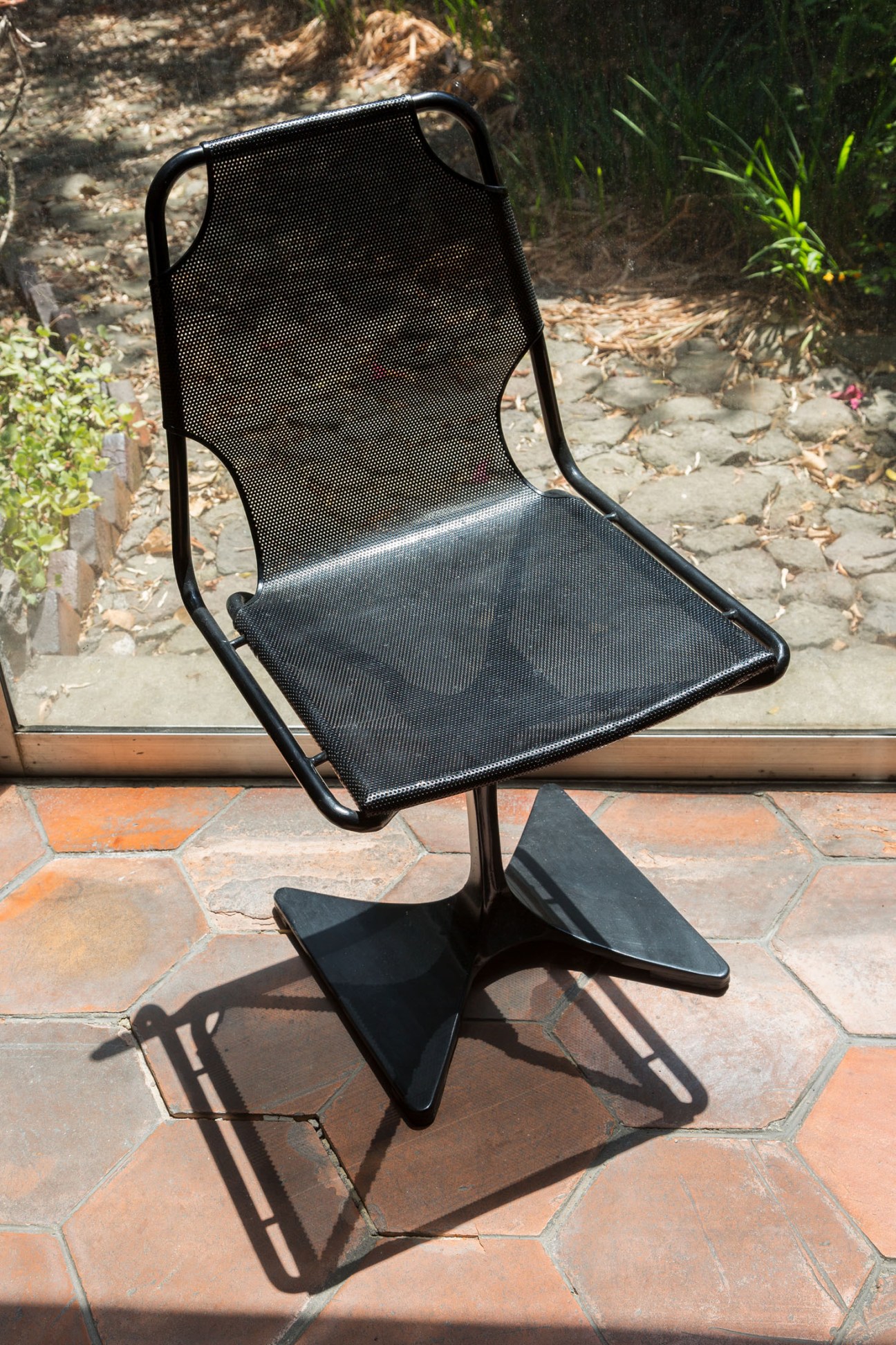 A black metal chair with perforated seat and back on a terracotta tiled patio with garden in background.