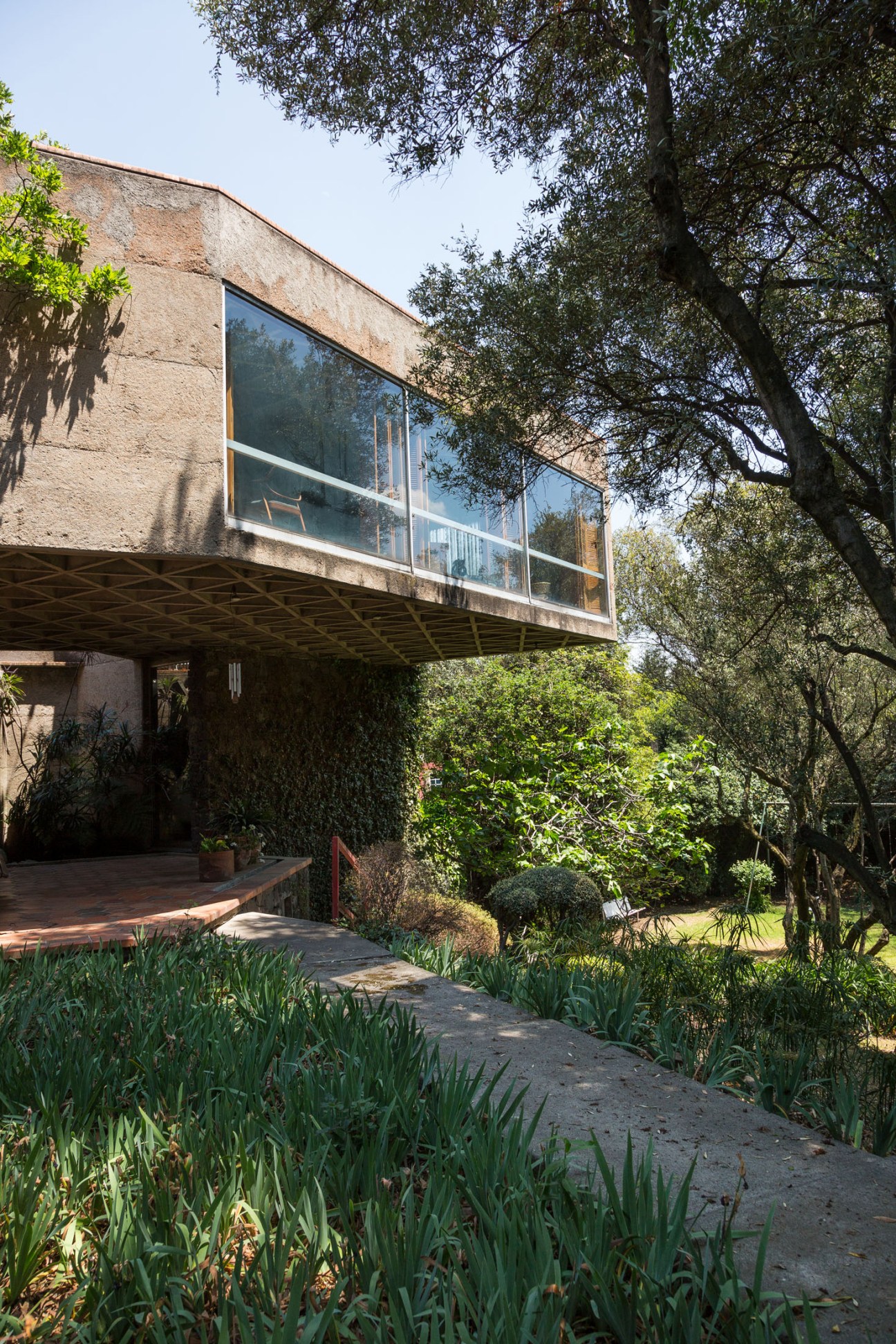 Concrete modernist house with large windows elevated on pillars, surrounded by lush gardens and trees.