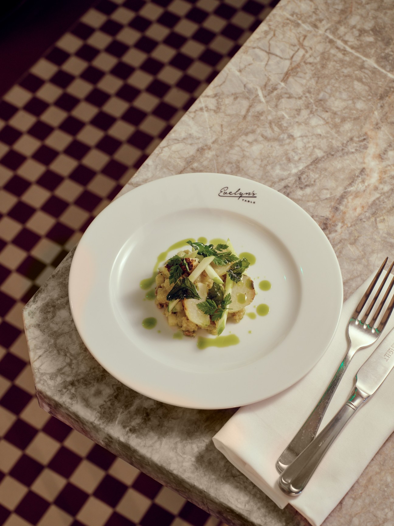 A plated dish at Evelyn's Table featuring vegetables and herbs with green sauce on a white plate.