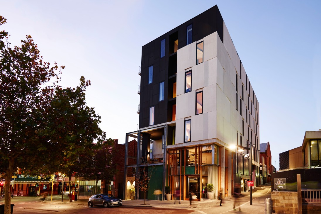 Modern multi-story hotel building with black upper facade and white lower section, illuminated ground floor entrance, city street at dusk.