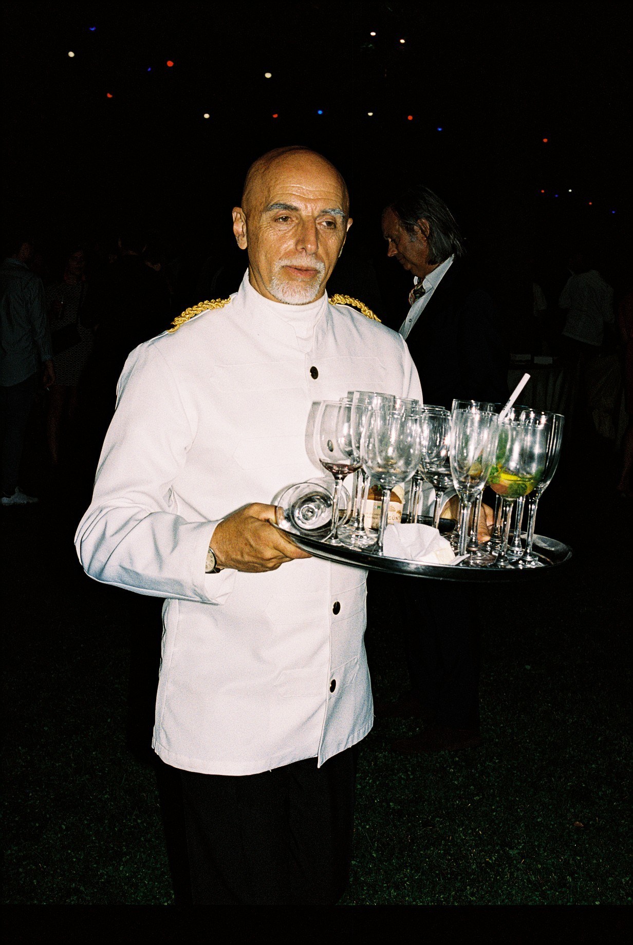 French ambassador Christian Masset in white dress uniform with gold epaulettes holding a tray of cocktails at a diplomatic reception.
