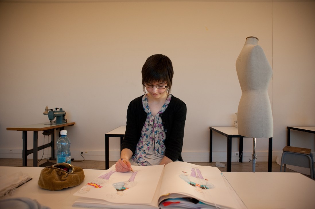 Fashion design student sketching designs at a table in studio with dress form and sewing machine nearby.