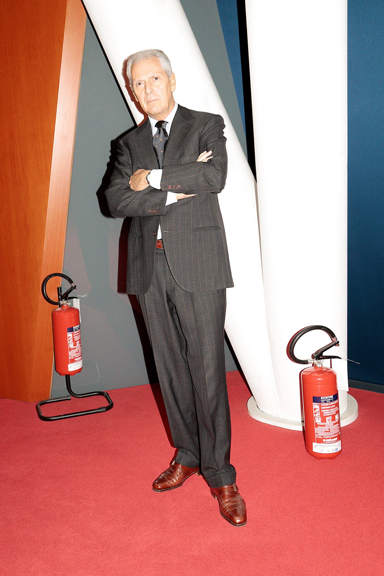 Pirelli CEO Marco Tronchetti Provera stands with arms crossed in a dark pinstriped suit against a geometric background with fire extinguishers.