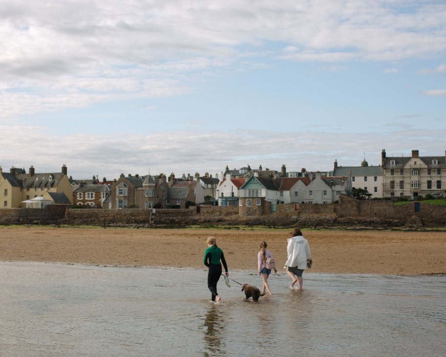 Three people and a dog wade in shallow water at a beach with a row of colorful stone cottages and buildings along the shoreline.