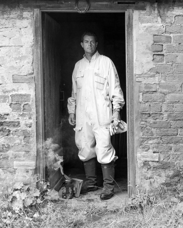 Man in white protective beekeeping suit standing in doorway of stone building.