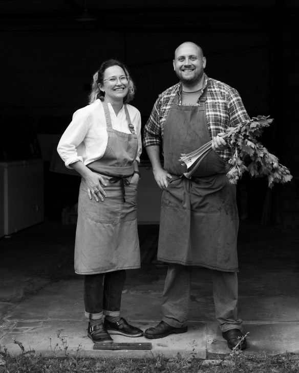Two people in aprons stand together holding fresh vegetables against a dark background.
