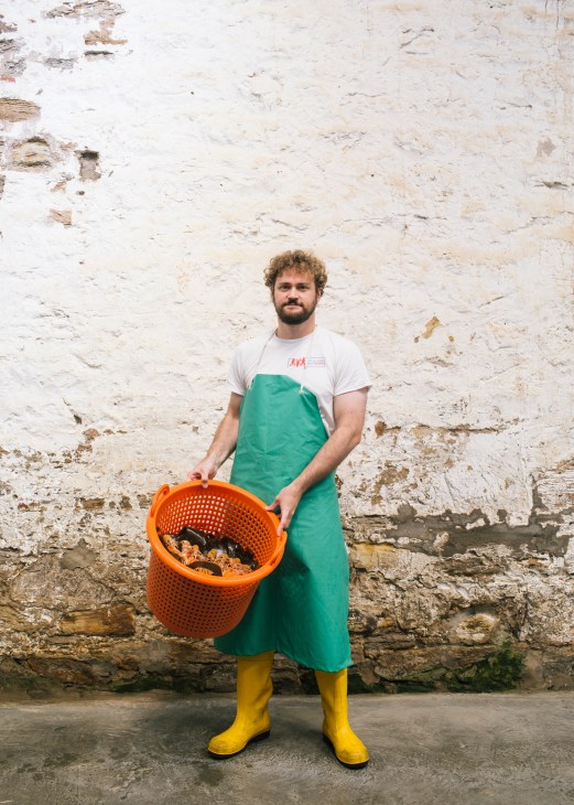Clement Boucherit of Langoustine the Box holds an orange basket of shellfish against a weathered stone wall.