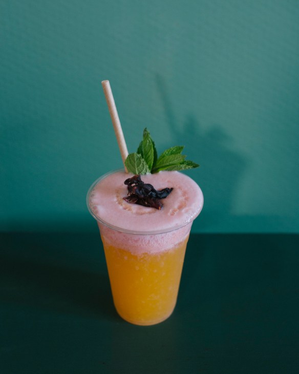 A layered drink with golden juice base and pink foam top, garnished with mint and dark edible flowers, served with a paper straw against a teal background.