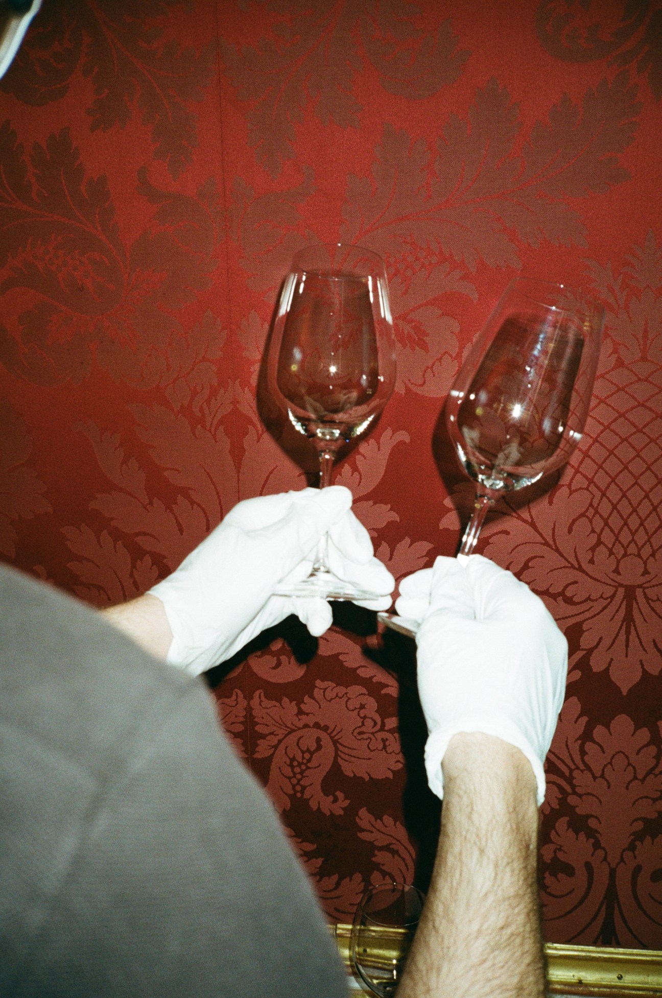 Two white-gloved hands holding wine glasses in a toast against an ornate red damask wallpaper background