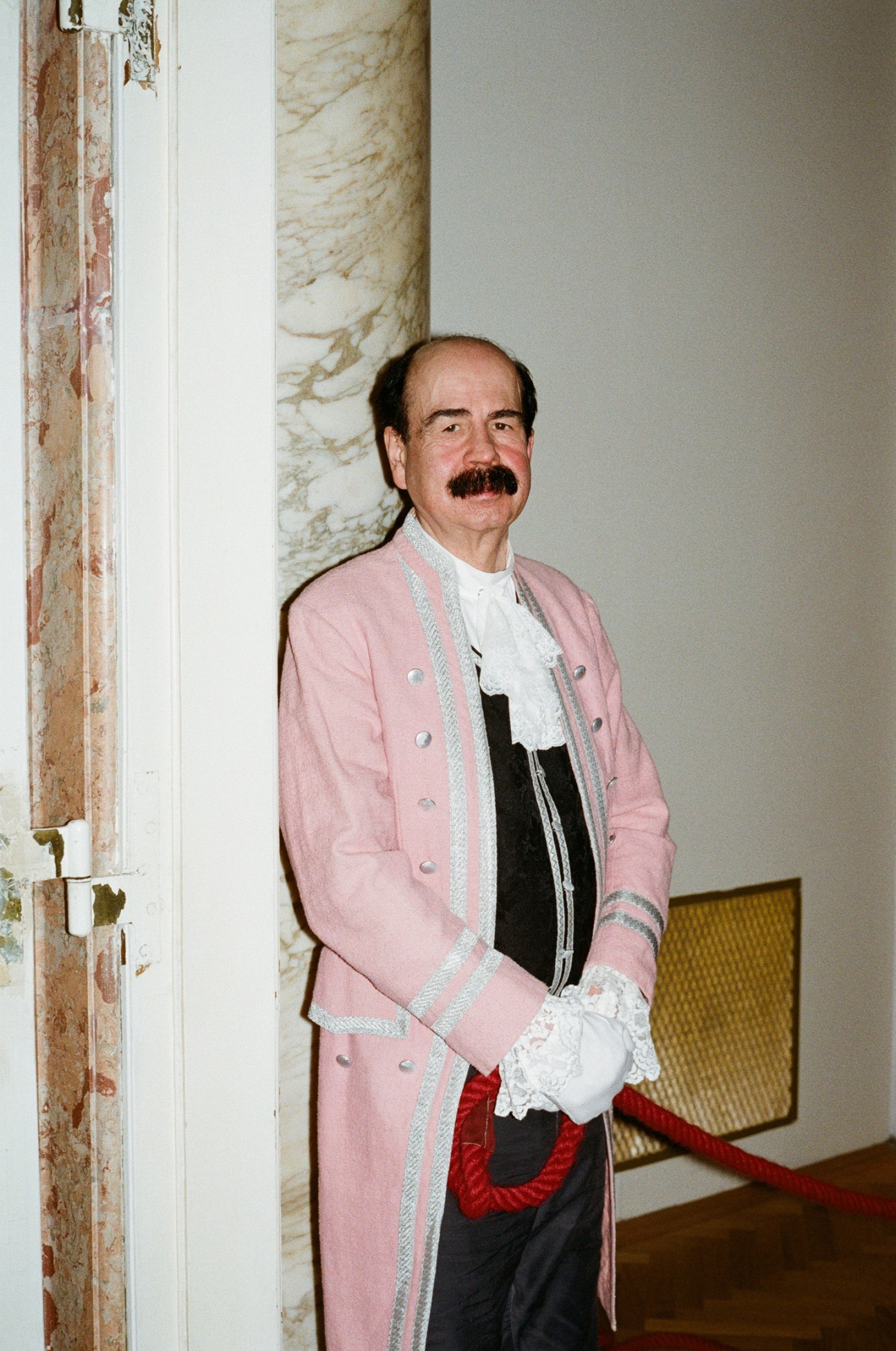 Man in pink 18th-century court attire with white ruffled shirt and red sash standing by marble column