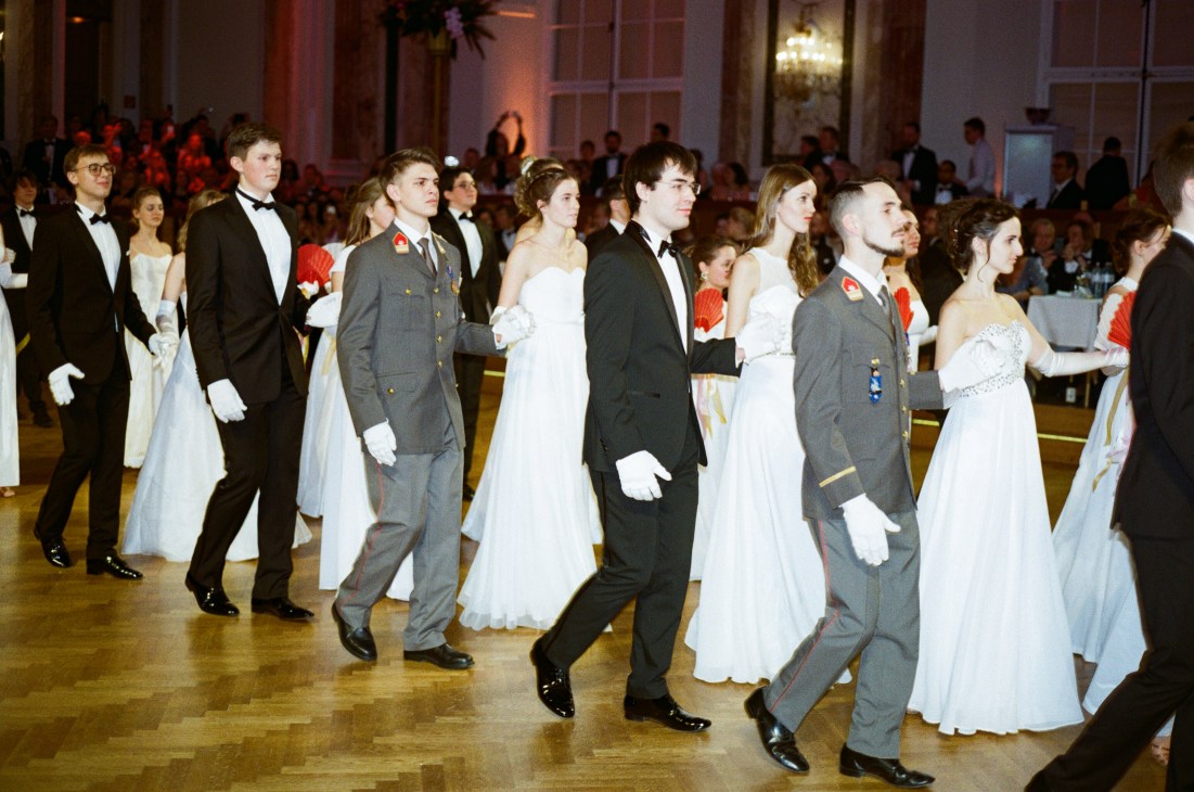 Young soldiers in grey military uniforms with red sashes dancing with women in white ball gowns at a formal Viennese ball