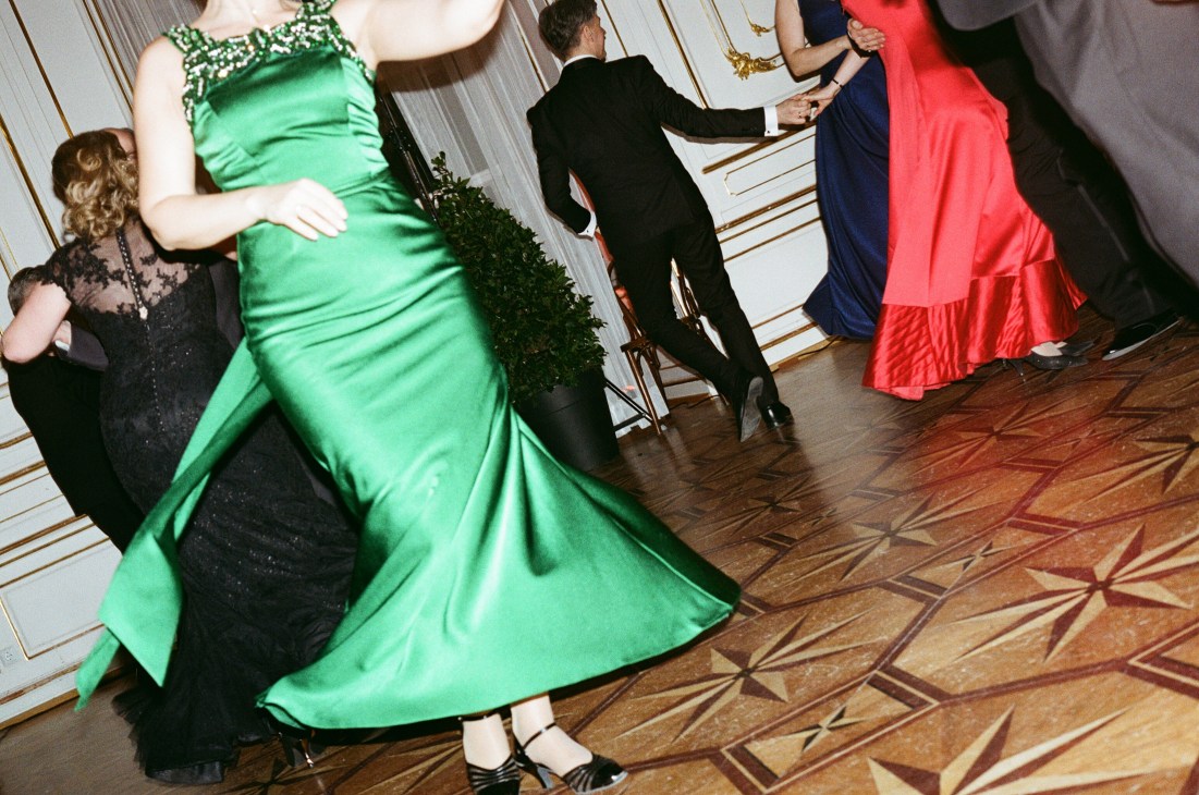 Ballgoers in formal evening wear dancing in an ornate ballroom with decorative wooden floors
