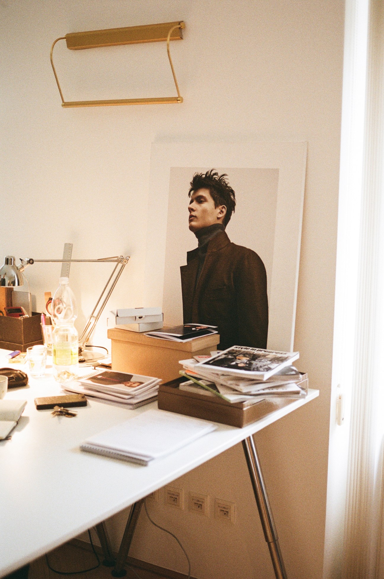 Man in dark turtleneck standing behind white desk with books and lamp in bright minimalist workspace