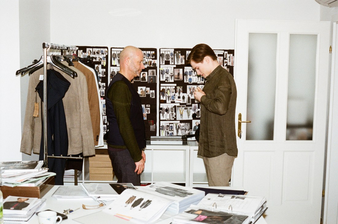 Two men in a design studio with clothing racks, mood boards, and fabric samples on a white table