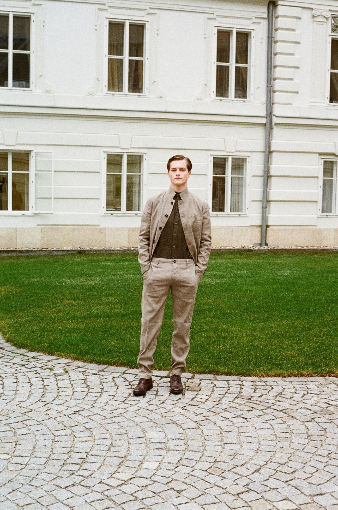 Man in beige suit standing on cobblestone street in front of white classical building