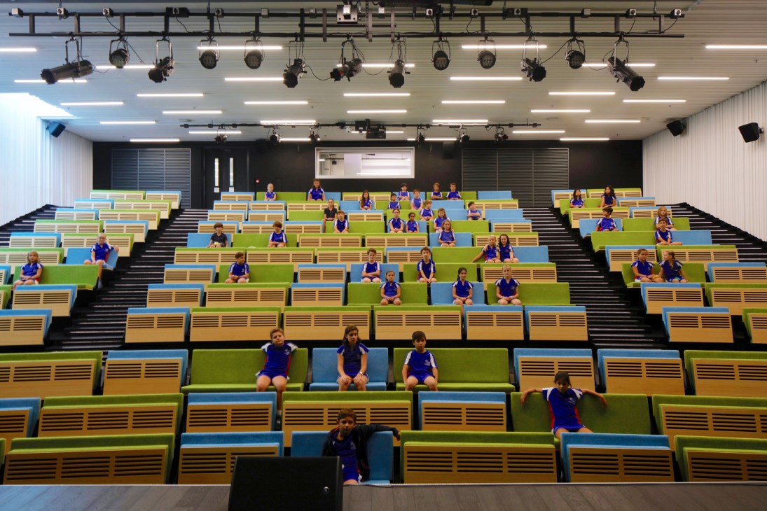 Students in blue uniforms sit in the 300-seat auditorium with colorful yellow, blue and green seating and overhead lighting rigs.