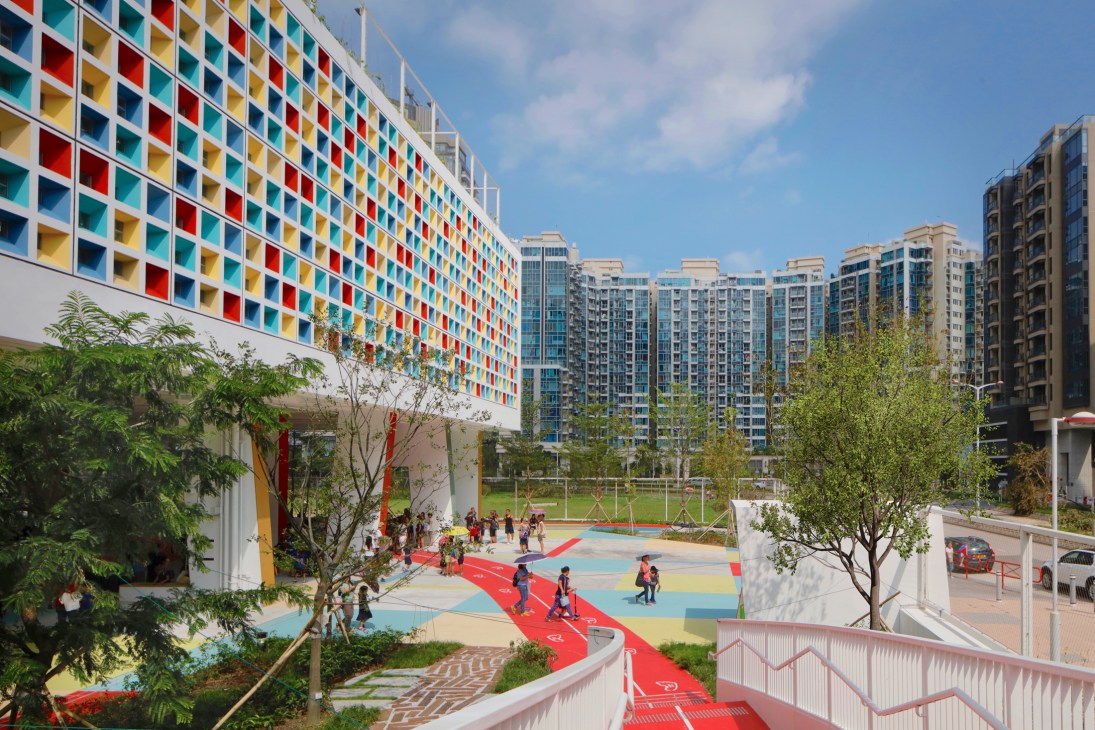 Elevated school building with colorful window facade overlooking a multicolored playground and sports court surrounded by tall residential towers.