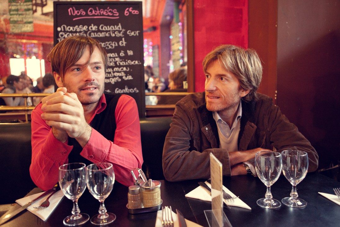 Two men dining at a restaurant with glasses of water and a menu visible in the warm-lit background.