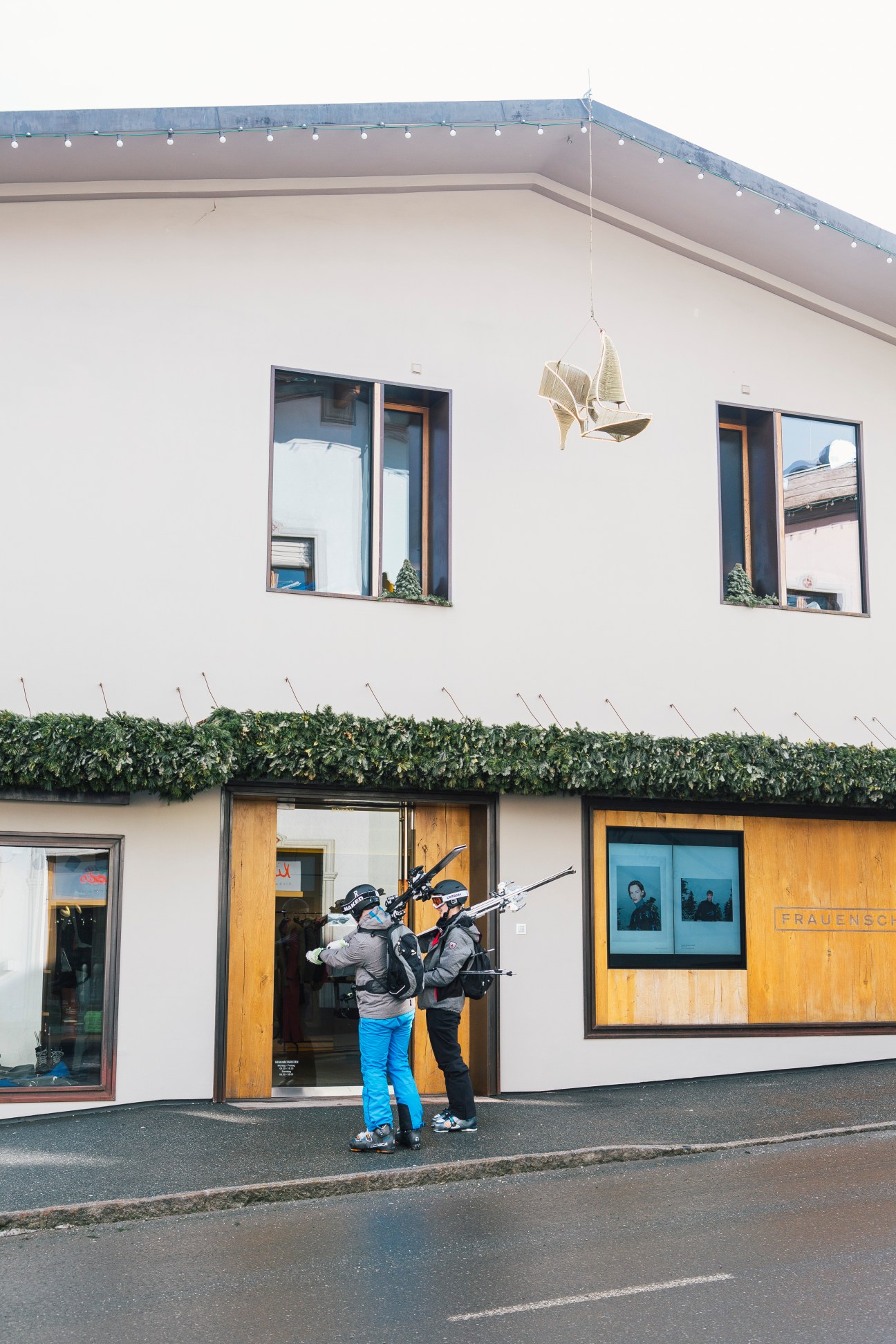Two people in winter ski gear carrying skis stand outside a modern building with wooden and glass storefronts