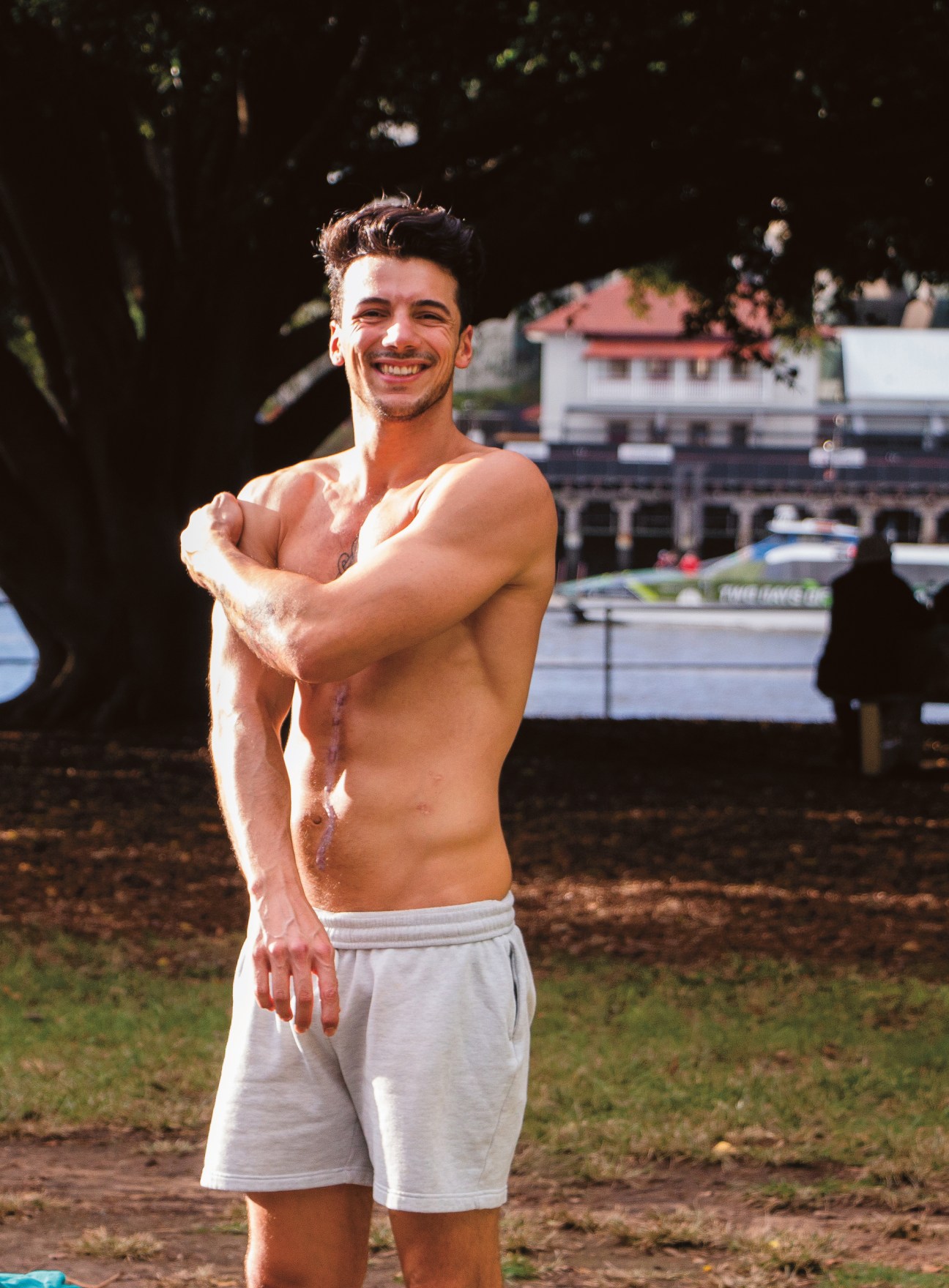 Shirtless man smiling at camera in sunny park overlooking Brisbane River and buildings.