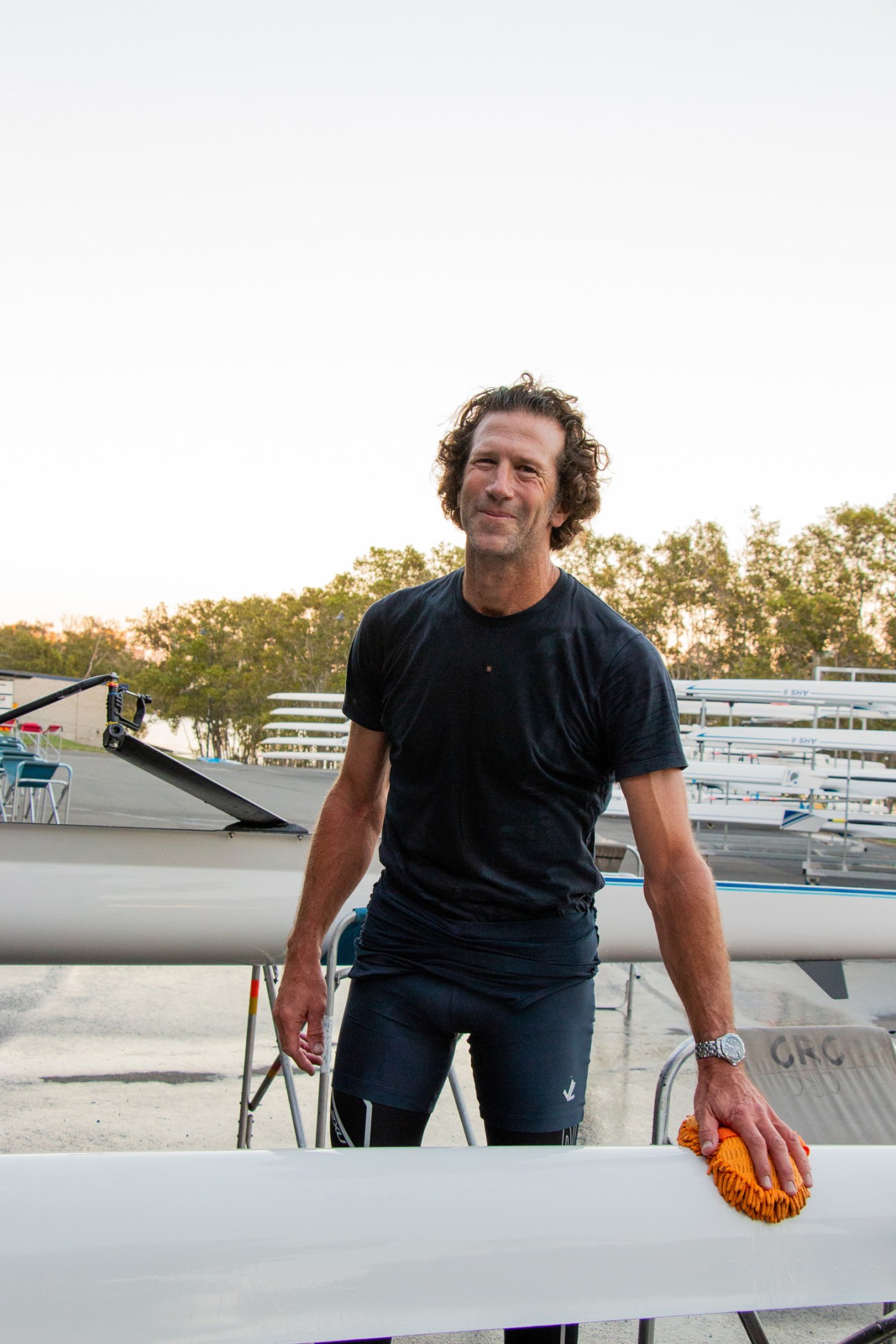 A man in a dark t-shirt stands at Commercial Rowing Club with rowing boats visible behind him.