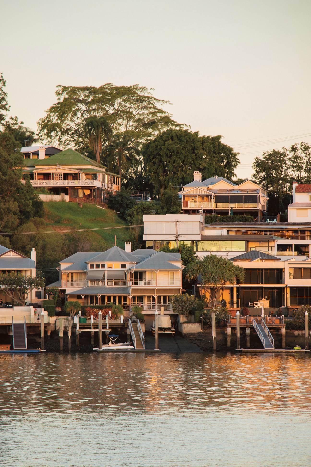 Waterfront houses with wooden decks and jetties along the Brisbane River at sunset.
