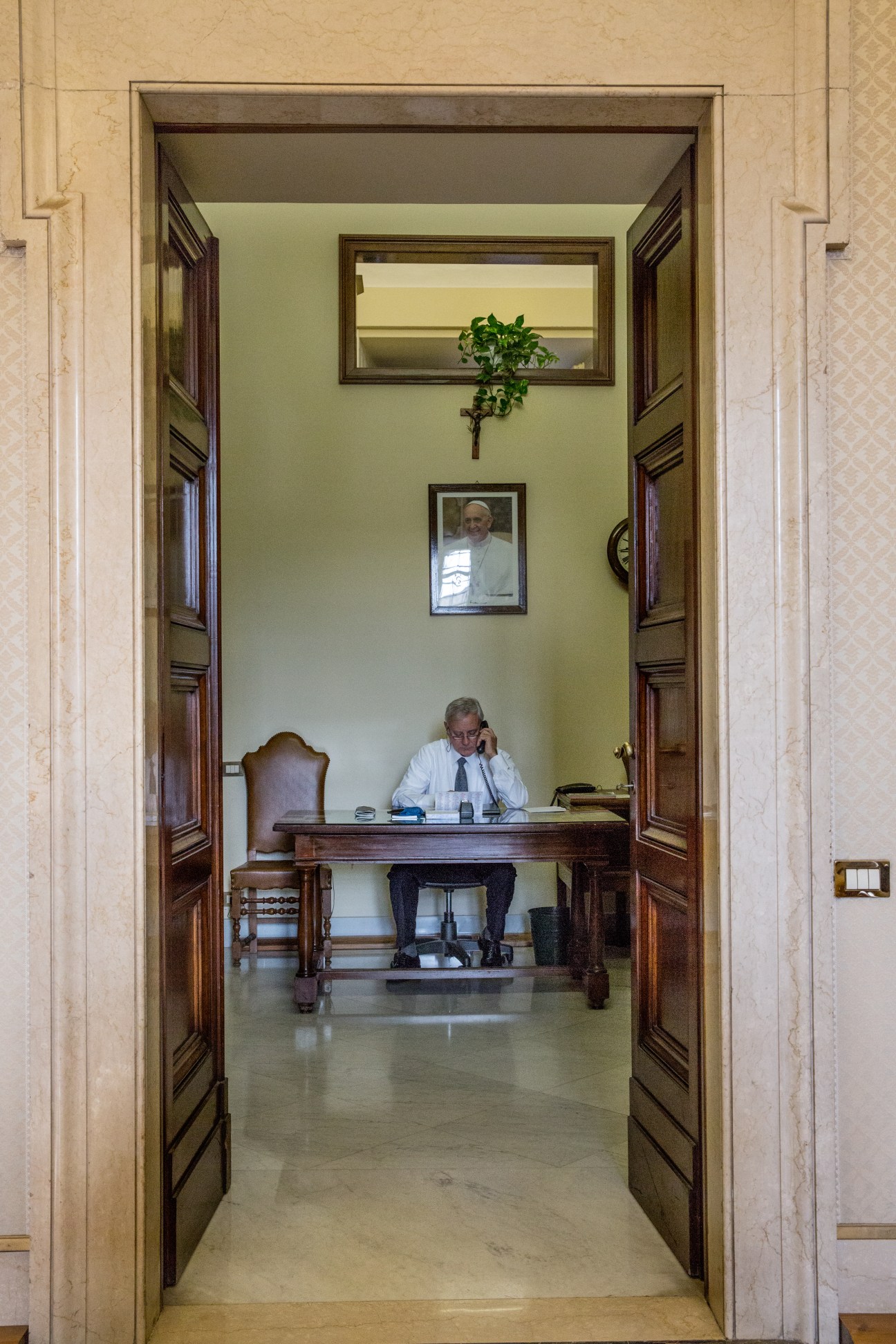 Man in white shirt and tie sits at wooden desk in pale green office room with portrait of Pope Francis on wall.