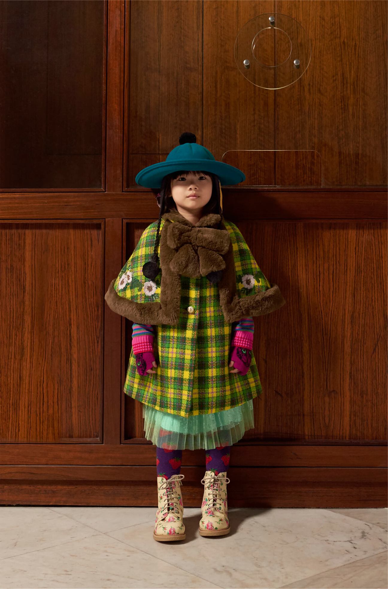 Child wearing colorful Gucci childrenswear including plaid coat, blue hat, and floral boots against wooden paneling