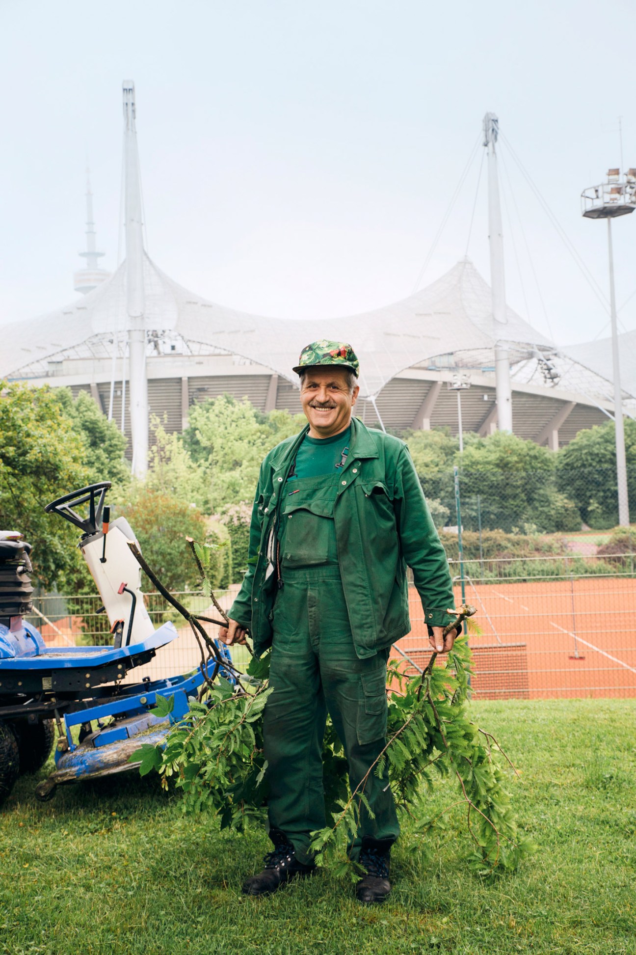 A gardener in green uniform and patterned cap holds pruning shears while standing in front of Munich's Olympic Stadium with its distinctive white tensile roof structures.