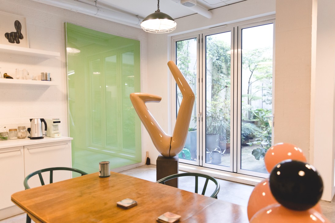 Modern kitchen with white cabinetry, green glass panel, wooden dining table, orange and black spheres, and a bronze abstract sculpture near garden doors.