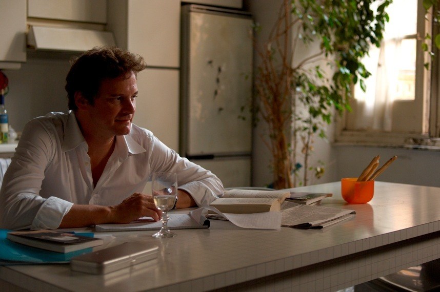 Man in white shirt sits at desk with glass of water and papers in bright room.