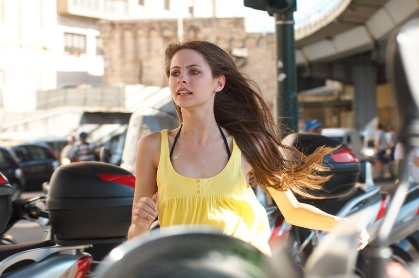 Woman in yellow top running through urban street with buildings and parked scooters in background