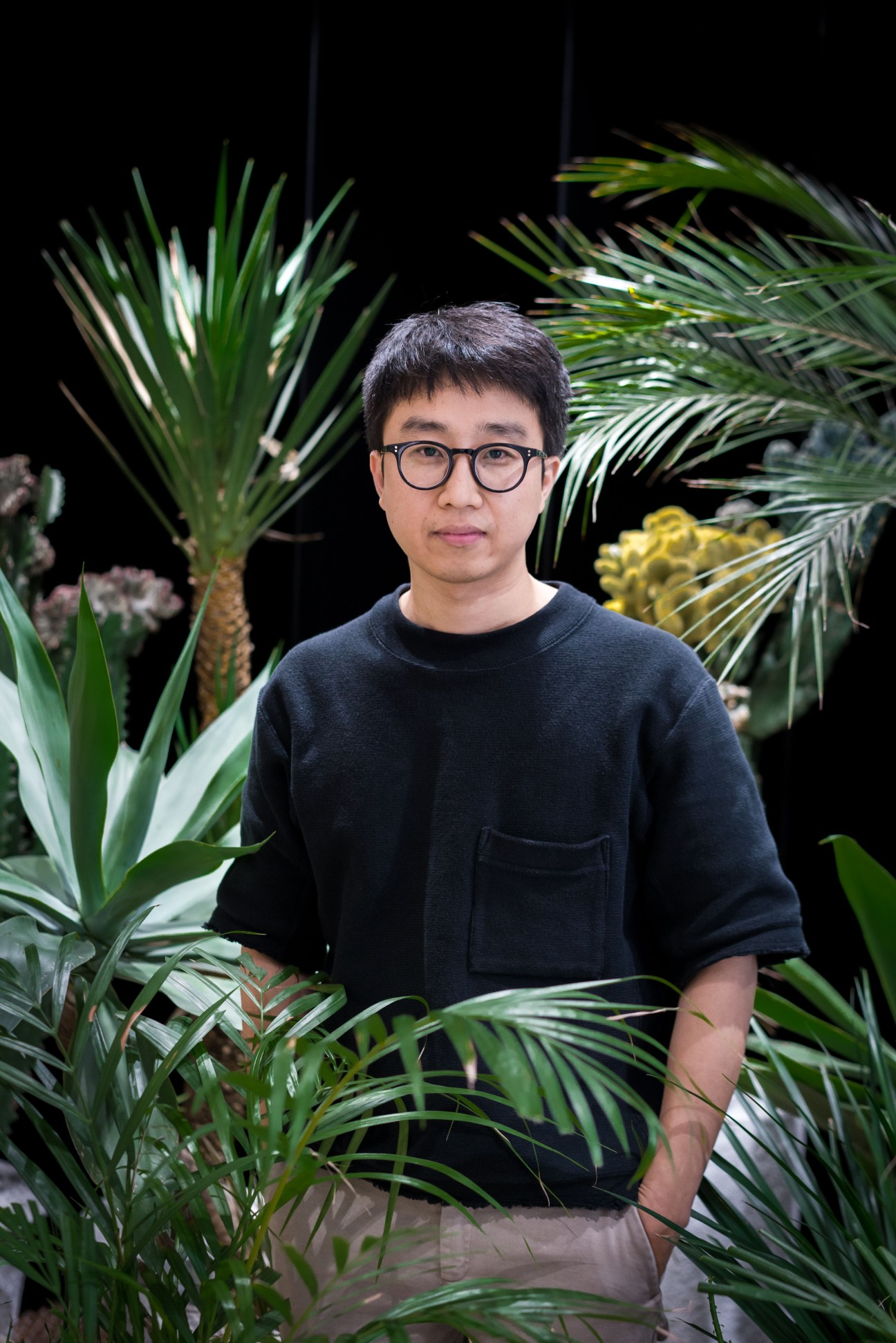 Hankook Kim, co-founder and CEO of Gentle Monster, wearing glasses and a black sweatshirt surrounded by tropical plants.